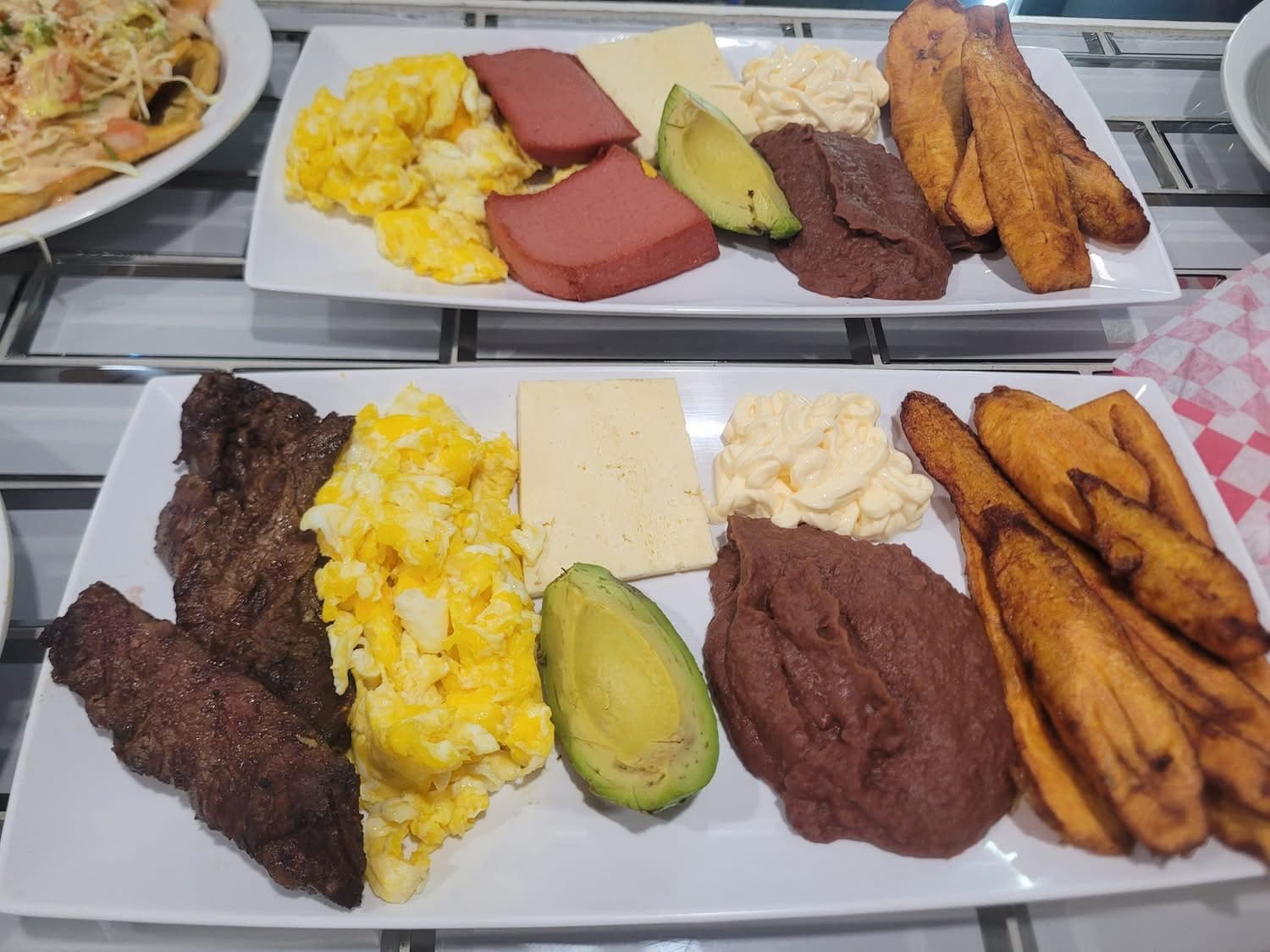 Two breakfast plates with scrambled eggs, steak, fried plantains, refried beans, avocado, and cheese.