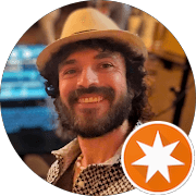 Smiling bearded man with curly hair in a tan hat and an orange star badge.
